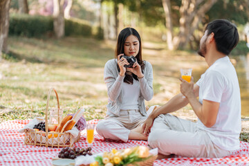 Happy asian couple travel. Woman taking photo of his boyfriend picnic time in park holidays vacation.