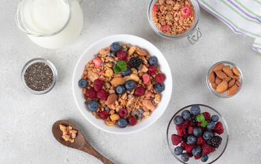 Crispy muesli with fresh berries, almonds in bowl and milk in jug, chia seeds for making pudding on light gray background