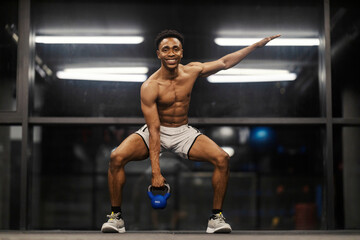 Strong muscular black sportsman in squatting position lifting kettlebell at gym.