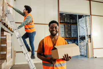 Obraz premium Portrait of diverse storage worker with box in hands smiling at camera while his coworker on ladder reaching for more boxes.
