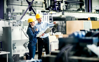 Two project managers standing in modern industrial factory, looking document in clipboard. Manufacturing facility with robotics, robotic arms and automation