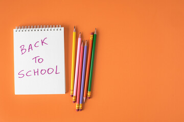 Close top down view of white spiraled notebook with back to school sign and colorful pencils on orange background with copy space