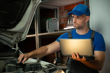 A mechanic in a blue shirt and cap stands in front of a car with the hood open. He holds a laptop in his hands and looks at the screen. There are tools and other equipment on shelves in the background