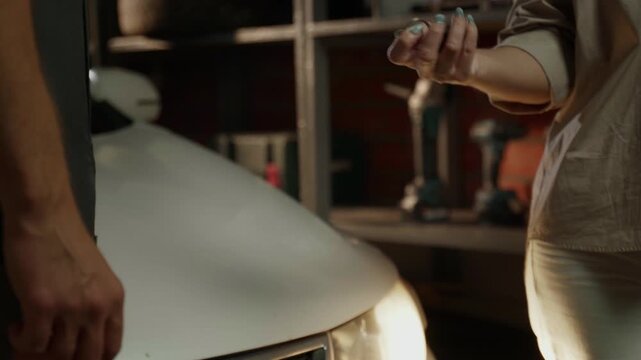 The mechanic hands over the car keys to the customer after the repair is completed in the garage. The mechanic is wearing a gray jumpsuit, and the client is wearing a beige shirt