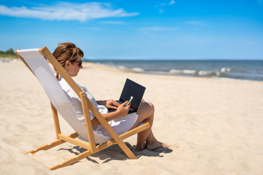 Remote work on beach. Young woman dressed in white dress and sunglasses sitting on deckchair and holding laptop and smartphone on white sandy beach by sea on sunny day. - Powered by Adobe