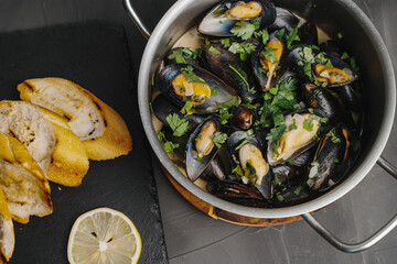 Mussels in white wine with parsley, toasted baguette slices and lemon on a gray table. A traditional dish of Mediterranean cuisine.