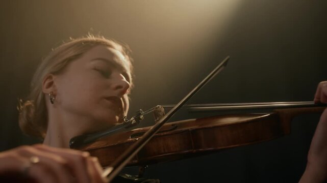 Chest up of beautiful young Caucasian woman playing violin while standing under projector light on black background