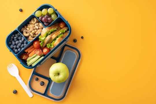 Healthy school snack in lunchbox with fresh fruits, vegetables, nuts, and sandwich arranged on yellow background
