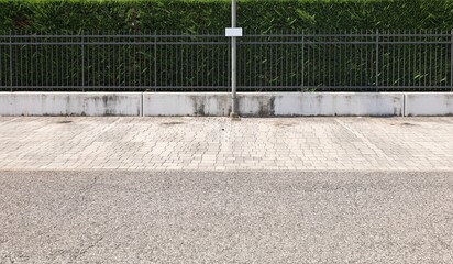 Pole on tiled sidewalk with a small blank sign hanging. Fence with metal black railing and evergreen privacy hedge on behind. Street in front. Background for copy space.