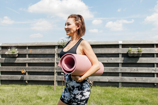 Smiling woman wearing sports clothing and holding exercise mat standing at back yard