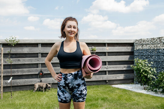 Mature woman holding exercise mat and standing with hand on hip in back yard