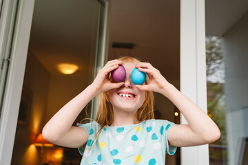 Happy girl covering eyes with colored easter eggs at home