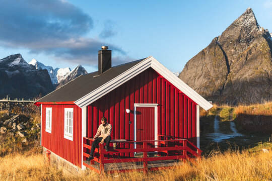 Man sitting on edge of railing in front of red log cabin at Nordic Islands