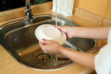 Asian woman washing dishes in the kitchen sink. scrubbing a plate with soapy sponge. Modern kitchen with wooden cabinets and stainless steel sink. Engaged in daily household chores,