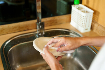 Beautiful Asian woman washing dishes in the kitchen. cleaning a plate under running water. Modern kitchen with wooden cabinets and stainless steel sink. Engaged in daily household chore.