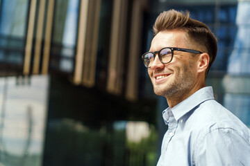Obraz premium Close-up of a smiling gentleman in glasses and a blue shirt, with a modern building in the background.