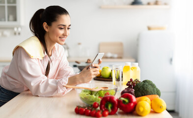 Smiling young woman using cellphone while cooking dinner in kitchen at home, cheerful casual female leaning on table, reading text messages on her cell phone and smiling, free copy space