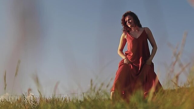 beautiful young long-haired brunette in long red summer dress erotical dances on green meadow at sunset in rays of sun in summer, slow motion