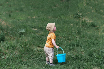 A little boy in a red T-shirt and a sand hat with a blue bucket stands on the green grass © Anastasiya Famina