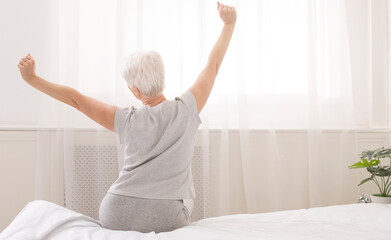 Senior woman sitting on her bed in morning, stretching with arms raised, back view, free space