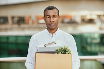A young black man in a white button-down shirt stands outside an office building holding a cardboard box filled with his belongings. He looks directly at the camera with a serious expression