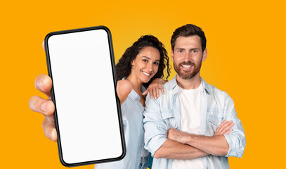 A couple stands in front of a bright yellow background. The woman has her arm around the man's shoulder and they both smile at the camera. The woman holds a smartphone with a blank screen
