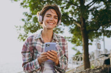 Fototapeta premium Young fun woman wear shirt casual clothes headphones hold use mobile cell phone listen music sits on bench rest in park near sea ocean beach outdoor seaside in summer day free time. Lifestyle concept.