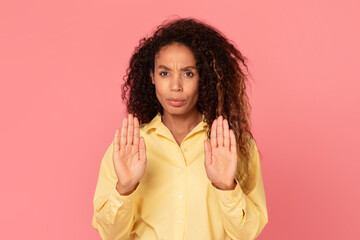 African American woman with curly hair is standing in front of a pink background, with both of her hands outstretched, palms forward, as if to say stop.