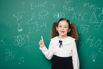 Little fun school girl 7-8 years old pony tails wear white shirt uniform point index finger indicate on area isolated on green blackboard background studio. Childhood kids education lifestyle concept.