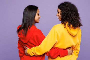 Rear view of two young fun little kid teen girls women they wearing red yellow hoodie casual clothes hug embrace put hands on back of each other isolated on plain purple background. Lifestyle concept.