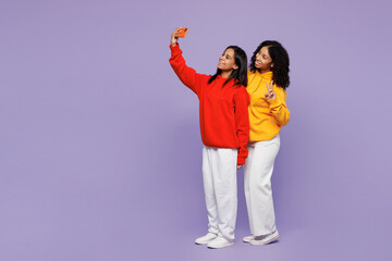 Full body two young little kid teen girls women they wearing red yellow hoodie casual clothes doing selfie shot on mobile cell phone show v-sign isolated on plain purple background. Lifestyle concept. © ViDi Studio
