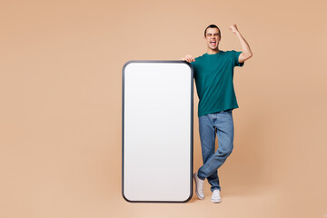 Full body young middle eastern man wear blue t-shirt casual clothes big huge blank screen mobile cell phone smartphone with workspace area do winner gesture isolated on plain pastel beige background