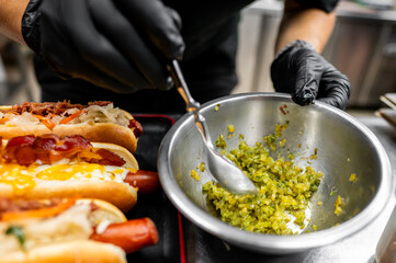 Close-up of hands in black gloves preparing hot dogs with bacon and cheese toppings, adding relish. Street food preparation, culinary concept. Perfect for food blogs, culinary websites