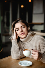 A woman is sitting at a table with a cup of coffee in front of her. She is wearing a sweater and she is resting her head on her hand. The scene suggests a quiet and relaxed atmosphere