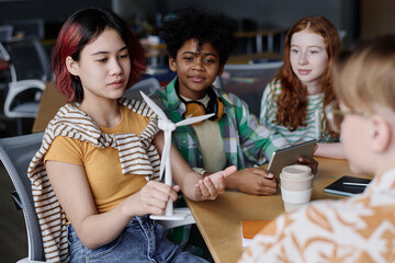 Asian girl holding wind turbine model discussing renewable energy sources with her classmates during Environmental Education lesson