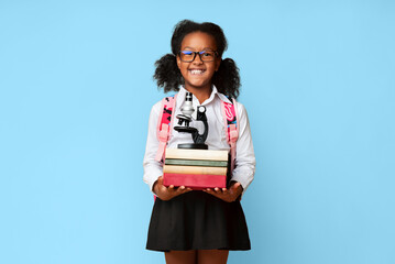 School Knowledge. Happy african girl holding books and microscope learning at home posing on yellow studio background.