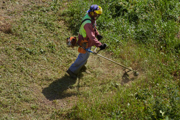 A gardener in overalls and protective overalls cuts overgrown grass with a lawnmower.