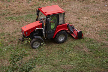 Obraz premium Red tractor with grass mower working in the field, grass mower in operation, harvesting grass with a mower on the tractor