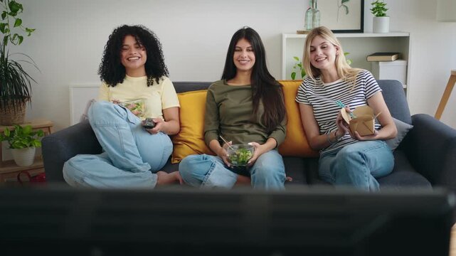Three flatmate girl friends having fun at home watching TV movie series show while eating healthy meal 