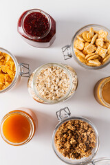 food storage and eating concept - close up of jars with oat, corn flakes, granola, cookies and spreads on white background