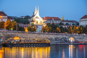 The Vltava River flows past the illuminated Emmaus Monastery in Prague, Czechia. The water reflects the lights of the city, creating a beautiful scene. A boat is docked near the riverbank.