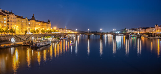 Obraz premium A view of the Rasin Embankment and Palacky Bridge in Prague, Czechia at dusk. The bridge is lit up, and the city lights reflect in the water. There are boats on the river, and the sky is a deep blue.