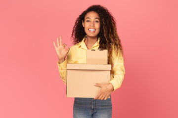 African American young woman with curly brown hair is standing in front of a pink background. She is holding two cardboard boxes and is making an okay sign with her left hand.