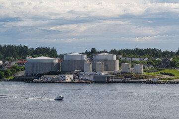 A small speedboat, speeding past a large industrial complex, on the coast of Norway