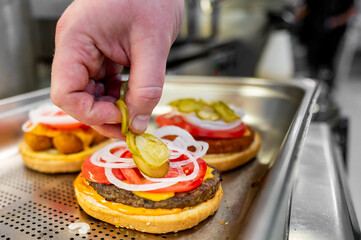 Close-up of a hand placing pickles on a freshly made burger with cheese, onions, and tomatoes. The background is blurred, emphasizing the burger assembly process. Ideal for food and culinary themes.