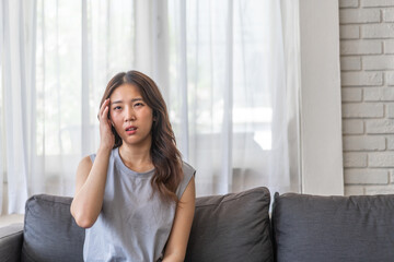 Young asian woman experiencing a headache sitting on a sofa with a pained expression, serious mood, health, wellness, stress, headaches and everyday struggles, pain and importance of self-care at home