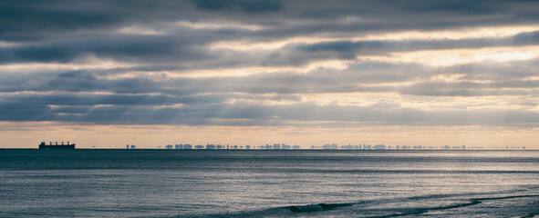 Fototapeta premium A lone ship sails on the calm ocean, with a distant city skyline visible on the horizon under a dramatic, cloudy sky.