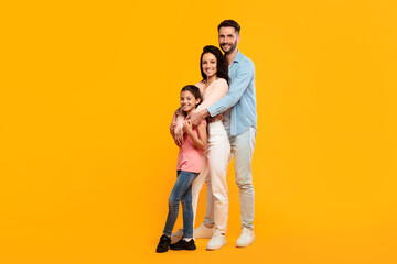 A family of three poses together against a bright yellow background. The father, mother, and young daughter are all smiling and looking at the camera