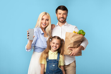 A happy family of three stands in front of a light blue background. The mother holds a smartphone in her right hand and smiles at the camera. The father holds a paper bag full of groceries