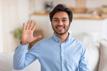 Arab man in a light blue shirt smiles and waves hello with his right hand, standing indoors in a home setting with a white couch partially visible in the background. © Prostock-studio
